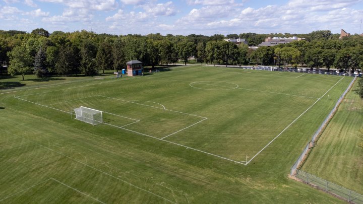 A wide aerial view of an empty soccer field with freshly mowed grass and clear white boundary lines. The field is surrounded by trees and a fence, with a small building located near the far corner. The goalposts stand ready at either end of the field, while the distant background shows parked cars and buildings.